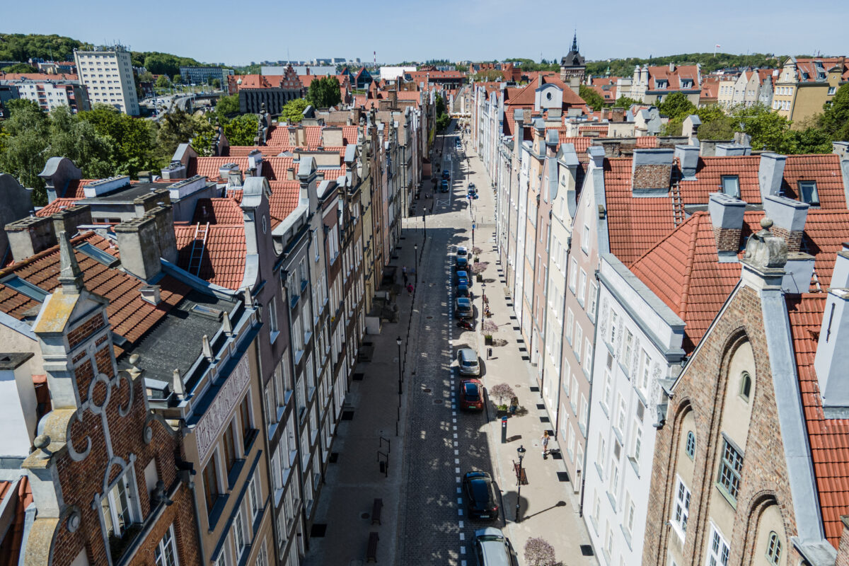 Z góry: długa, brukowana Ulica Ogarna. Kolorowe kamienice z czerwonymi dachami, zaparkowane auta, piesi na chodnikach.