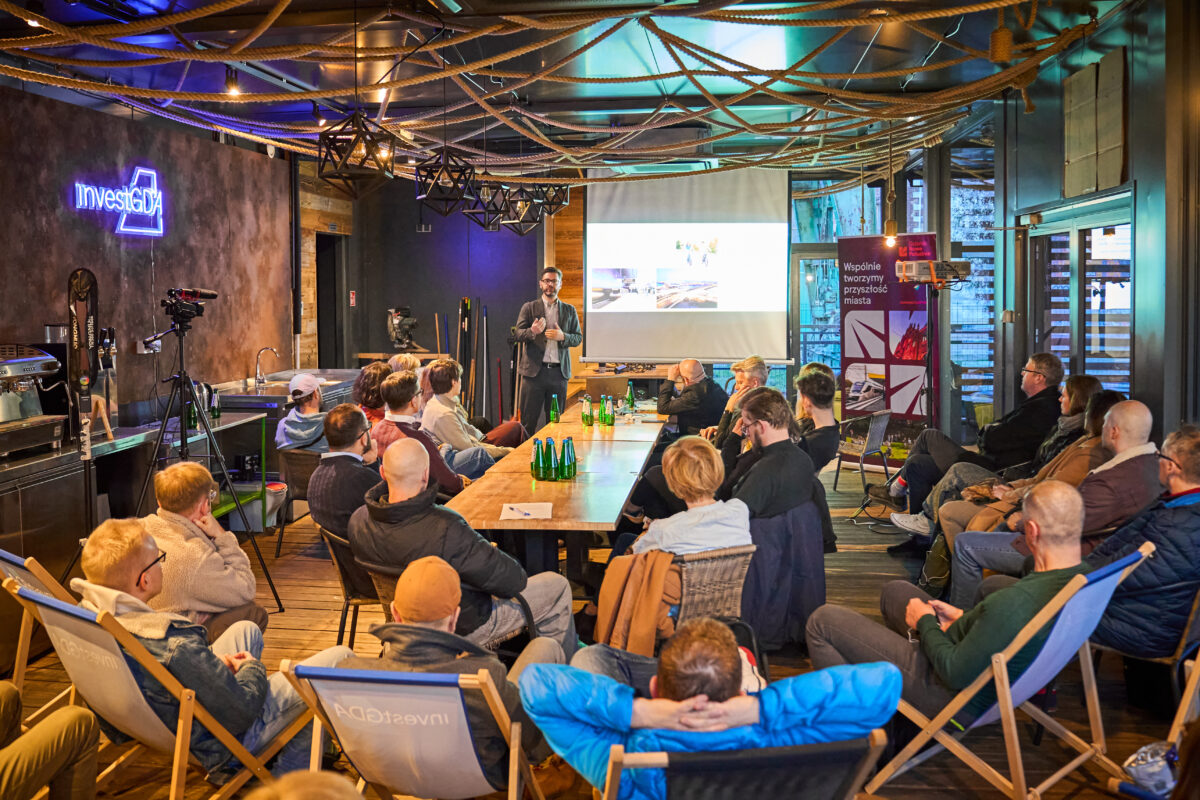 Mężczyzna prowadzi prezentację dla grupy ludzi siedzących w nowoczesnej sali z projektorem i neonem "InvestGDA".