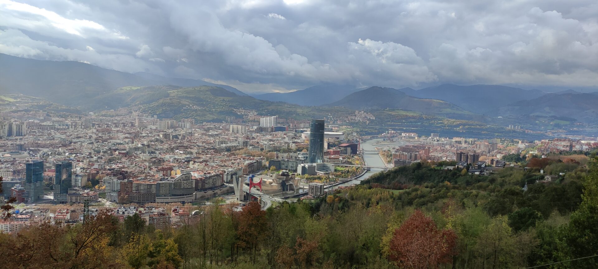 Panorama Bilbao: rzeka Nervión, Muzeum Guggenheima, wieżowiec, miasto otoczone górami pod zachmurzonym niebem.