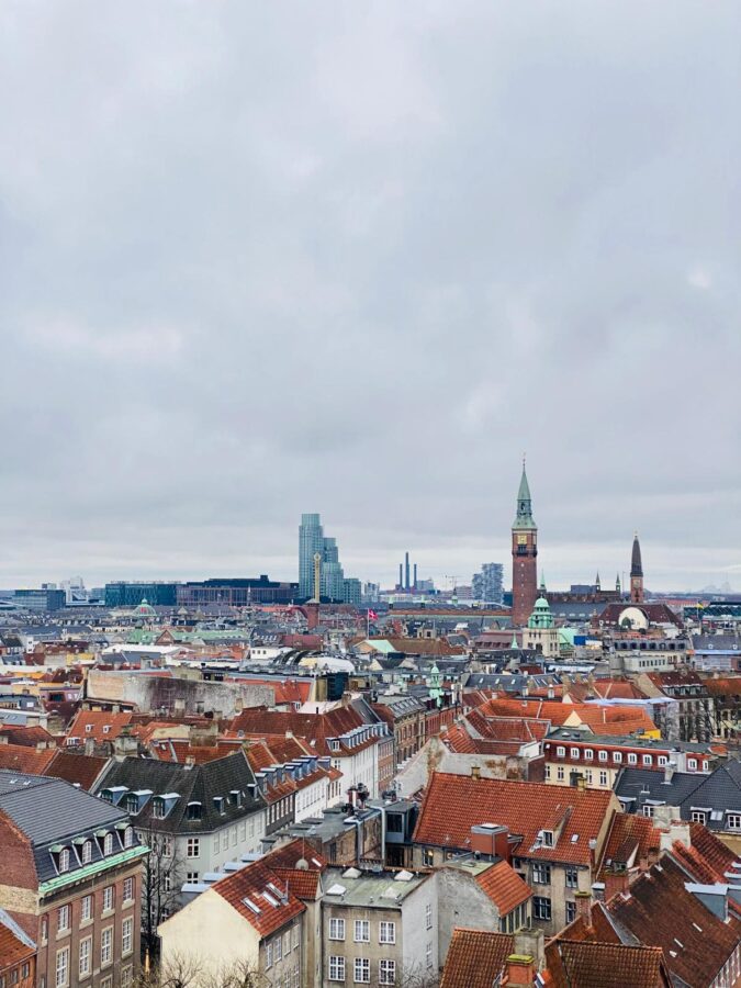 Panorama miasta: gęsta zabudowa z czerwonymi dachami, wieża zegarowa, nowoczesny wieżowiec. Pochmurny dzień.